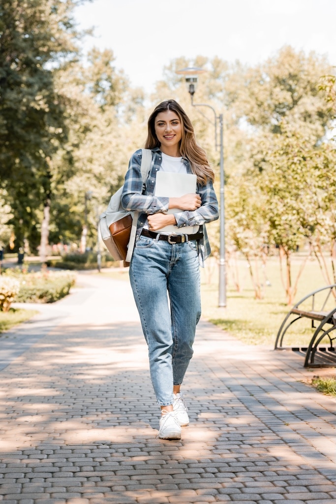 woman in plaid shirt holding laptop and looking at camera while walking in park